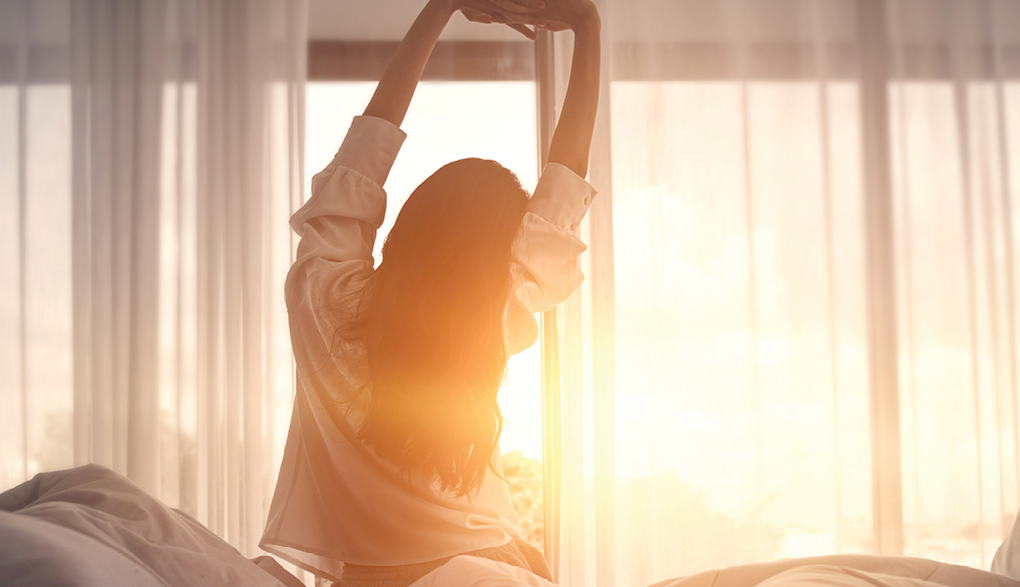 A woman stretching in the morning sunlight by a window, symbolizing the importance of arriving early to fully enjoy and immerse yourself in local folk festivals.